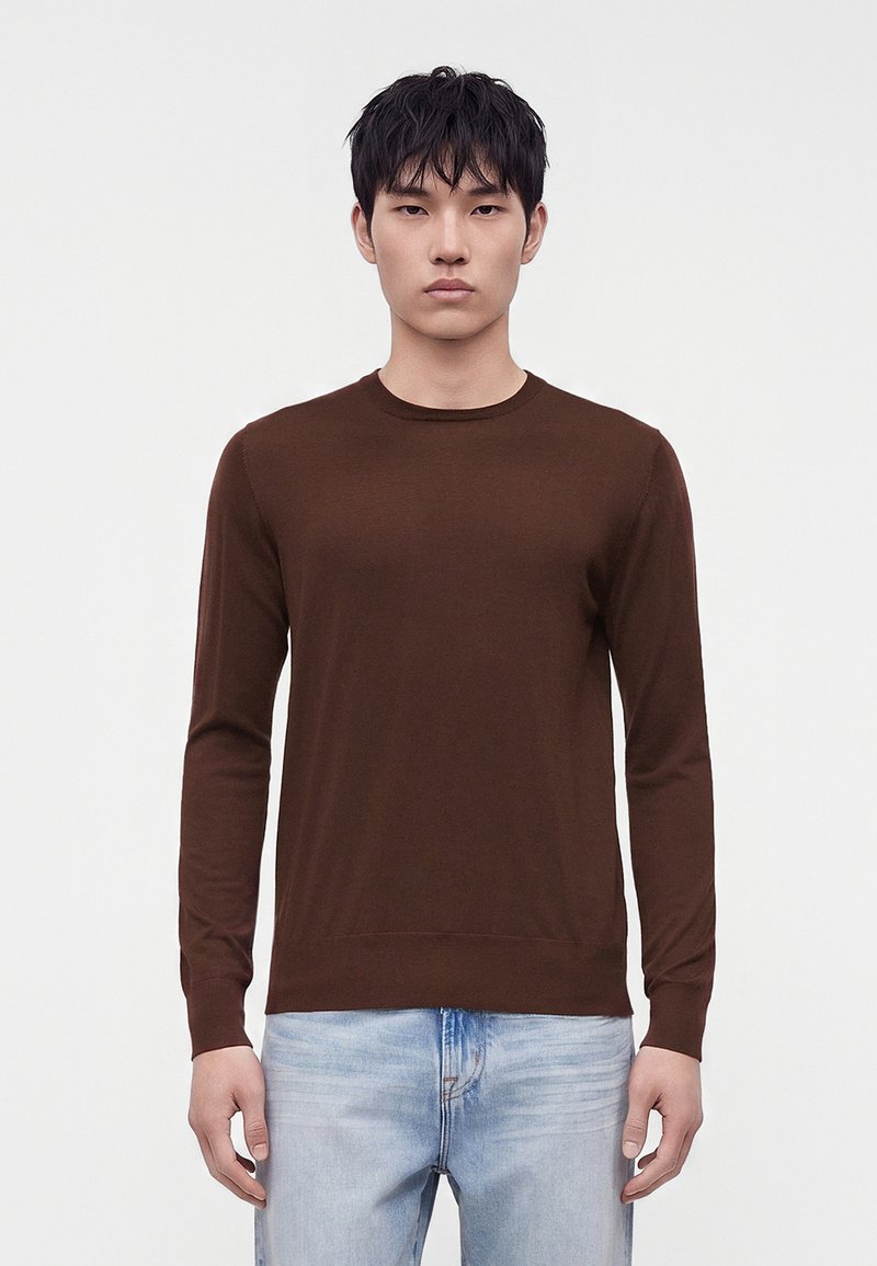 Jeune homme aux cheveux noirs portant un pull marron uni à manches longues et un jean bleu clair, debout sur un fond blanc.