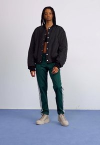 Giubbotto bomber nero con bottoni a pressione bianchi, pantaloni della tuta verdi con strisce bianche e sneaker beige indossati da un modello su uno sfondo neutro.