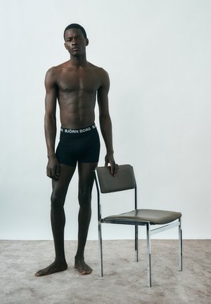 Λεπτός άνδρας με μαύρα εσώρουχα Björn Borg στέκεται ξυπόλητος πάνω σε χαλί, ακουμπώντας το χέρι του σε καρέκλα με πλαίσιο από χρώμιο και γκρι κάθισμα και πλάτη.