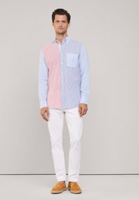 Camisa de botones a rayas con un diseño dividido: una mitad roja y la otra azul. Presenta un bolsillo en el pecho. Combinada con pantalones blancos y zapatos marrones.
