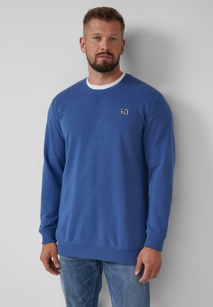Man with beard wearing blue long-sleeve crewneck sweatshirt and light blue jeans, standing against a plain white wall.