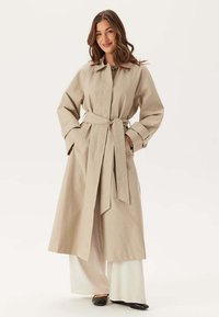 Beige trenchcoat med bälte, stor krage och vida ärmar. Bärs över vita vida byxor. Slät textur, minimalistisk design.