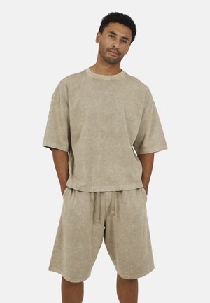 Mężczyzna stojący z rękami w kieszeniach, ubrany w luźną, beżową, wypraną koszulę z krótkim rękawem oraz pasujące szorty ze ściągaczem na tle białego tła.