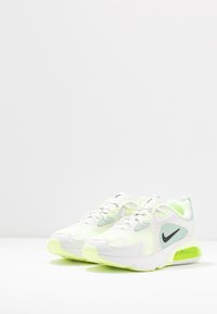 Zapatillas deportivas blancas con superposiciones de malla, acentos verde lima y el logotipo de Nike en negro. Presentan una amortiguación visible y suelas texturizadas.