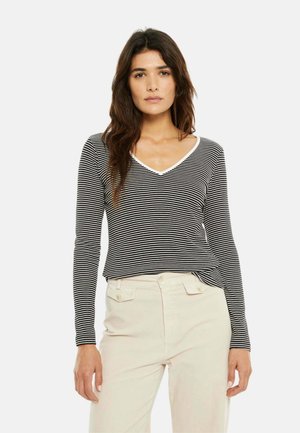 Mujer con cabello largo y oscuro que lleva una blusa de manga larga a rayas negras y blancas y pantalones de talle alto color beige claro, de pie frente a un fondo blanco.