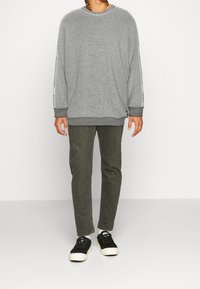 Persona in piedi con un maglione grigio texturizzato con lettering sulle maniche, indossando pantaloni verde oliva e sneaker nere su uno sfondo bianco.