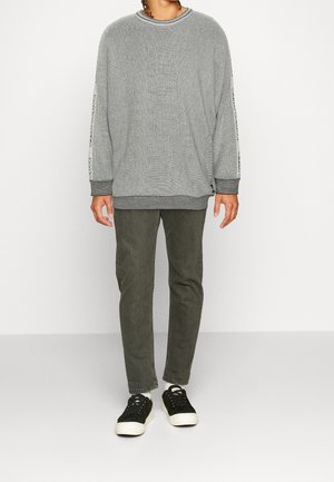 Persona de pie con un suéter gris texturizado con letras en las mangas, usando pantalones verde oliva y zapatillas negras frente a un fondo blanco.