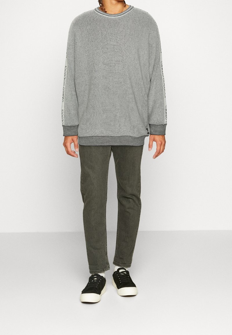 Persona in piedi con un maglione grigio texturizzato con lettering sulle maniche, indossando pantaloni verde oliva e sneaker nere su uno sfondo bianco.