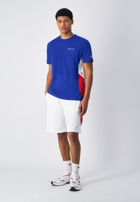 Blaues T-Shirt mit kurzen Ärmeln und roten sowie weißen seitlichen Akzenten und Logo. Kombiniert mit weißen Shorts und weißen Sneakers mit roten Highlights.