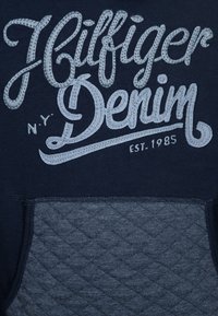 Felpa navy con logo grigio ricamato "Hilfiger Denim." Presenta una tasca testurizzata a forma di cucitura nella parte inferiore, tessuto morbido e scollatura a girocollo.