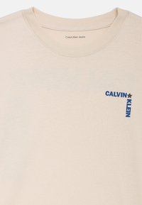 Světle béžové bavlněné tričko s kulatým výstřihem. Na levé části hrudi se nachází modrý potištěný logo s nápisem "CALVIN KLEIN". Měkká textura.