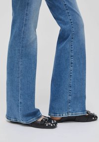 Jeans en denim bleu évasés avec un lavage clair, associés à des chaussures plates noires ornées de détails métalliques et de boucles sur les orteils.