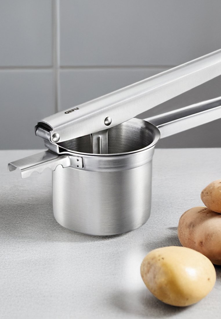 Edelstahl-Kartoffelstampfer mit langen Griffen, einer Schüssel zum Stampfen und einem Hebelmechanismus; positioniert in der Nähe mehrerer ungeschälter Kartoffeln.