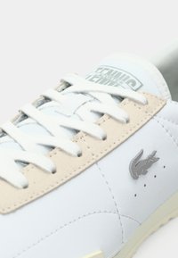 Sapatilha de couro branco com acabamentos em camurça bege, bico redondo, atacadores brancos, logo em crocodilo prateado e detalhes de perfuração na lateral.