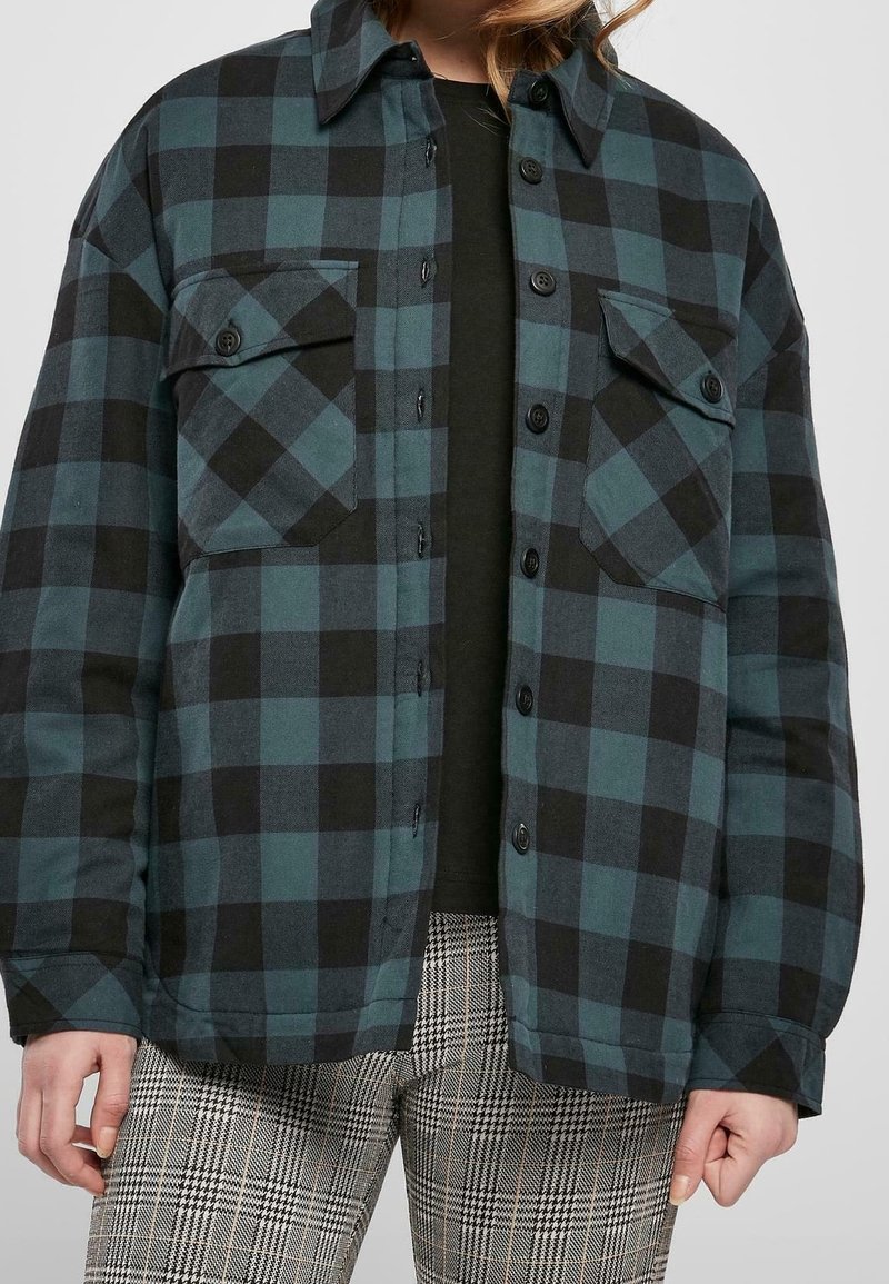 Personne portant une veste à carreaux bleu sarcelle et noir à boutons sur une chemise noire et un pantalon à carreaux gris, montrée des épaules à la mi-cuisse.