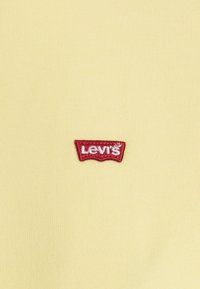 Tela de algodón amarilla con un logo de Levi's bordado en rojo en un acento con forma de etiqueta. La textura parece suave y ajustada.