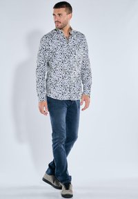 Weißes Langarmhemd mit grünem botanischem Muster, kombiniert mit dunkelblauen Jeans und grauen Sneakers. Das Model steht vor einem schlichten Hintergrund.