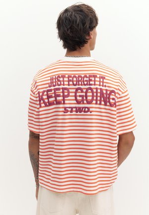 Homme aux cheveux foncés portant un t-shirt rayé orange et blanc avec l'inscription « JUST FORGET IT, KEEP GOING STWD. » en grandes lettres violettes dans le dos.