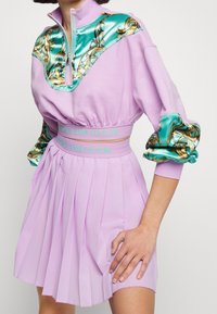 Lavendelfärgad sweatshirtklänning med dragkedja i halsen och satinäckenter i turkos med ett guldmönster. Plisserad kjoldetalj och varumärkesmidja.