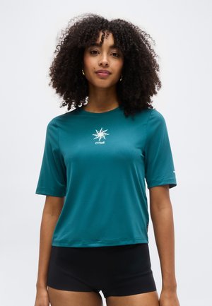 Göndör hajú nő, aki kékeszöld O'Neill rövid ujjú felsőt és fekete rövidnadrágot visel, egyszerű fehér háttér előtt áll.