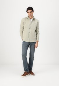 Cămașă butonată de culoare verde deschis, cu buzunar pe piept, purtată cu blugi albaștri și adidași maro. Textură netedă a țesăturii, design casual.