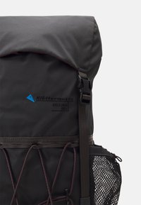 Black backpack with a roll-top closure, reinforced seams, adjustable straps, mesh side pocket, and contrasting red accents. 25L capacity.