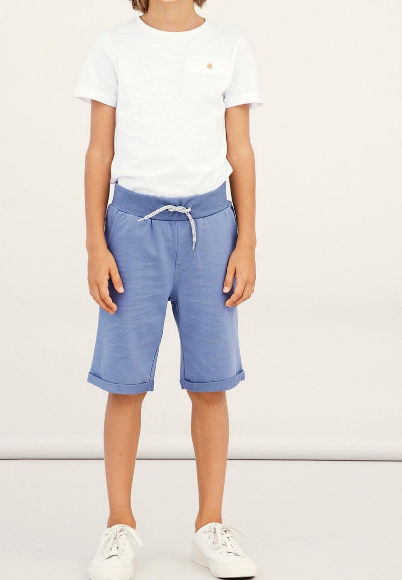 Enfant portant un t-shirt blanc à manches courtes, un short bleu mi-long avec cordon et des baskets blanches, debout devant un fond uni.