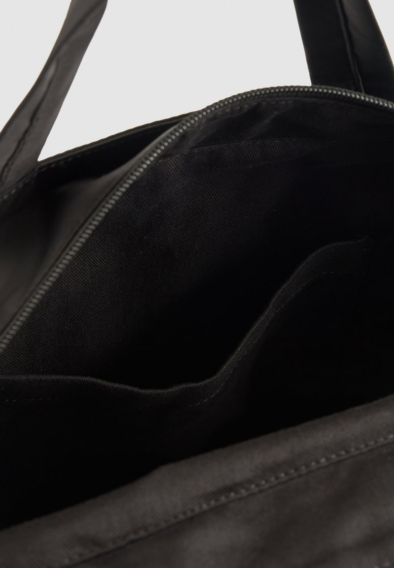 Interior de bolsa tote negra, hecha de tela suave con cierre de cremallera. Presenta un único compartimento y un bolsillo deslizante adicional en el interior.