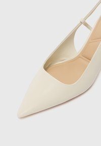 Spiss tå høyhælte sko i kremfarget lær med utskjæringsdesign på siden og en glatt, blank overflate. Innvendig har den en beige fôr.