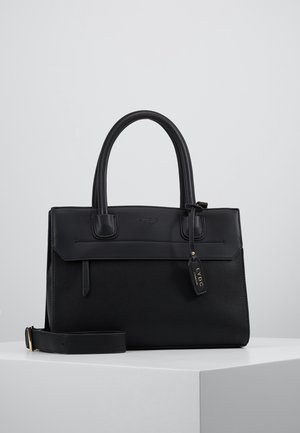 Sac à main structuré noir avec doubles poignées, bandoulière amovible, logo discret de la marque et une petite étiquette suspendue sur une surface blanche.