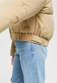 Chaqueta acolchada marrón claro con dobladillo elástico y detalles de botones, usada sobre unos vaqueros azules desgastados, mostrando una textura de tela lisa.