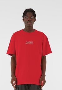 Punane suurendatud T-särk, mis on valmistatud puuvillast, millel on valge graafiline logo rinnal, lühikesed varrukad ja ümar kaelus.