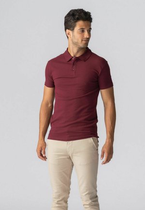 Mand iført en tætsiddende bordeaux polo shirt og beige bukser, stående og kiggende til sin venstre side mod en ensartet, lys grå baggrund.
