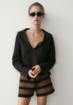 Femme portant un haut noir à manches longues avec col en V, des lunettes de soleil noires et un short en maille rayé marron et noir, debout contre un mur uni.