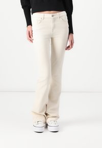 Jean évasé beige en denim, avec une fermeture à boutons devant, cinq poches et une texture lisse. Associé à des baskets blanches.