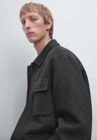 Donker wollen jack met een kraag, een enkele zak en een strak ontwerp. De stof lijkt gestructureerd met een zachte afwerking, geschikt voor koelere weersomstandigheden.