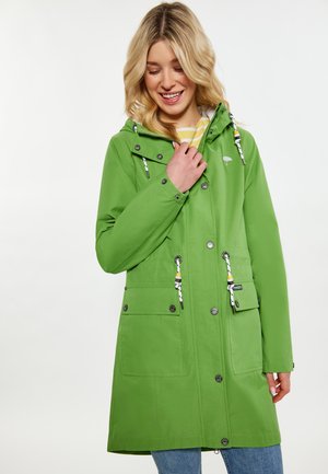 Donna che indossa un impermeabile verde con cappuccio, dettagli con bottoni e coulisse, sorridente mentre sistema il colletto.