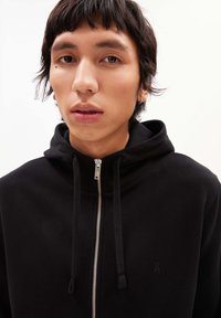 Felpa nera con zip realizzata in materiale morbido, dotata di colletto alto, cordini e un logo discreto vicino alla zip. Texture liscia.