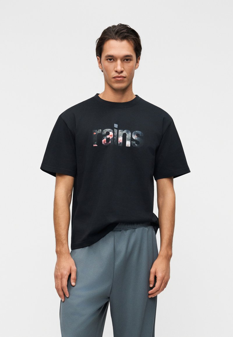 Νέος άνδρας με μαύρο μπλουζάκι με λουλουδάτο κείμενο "rains" και χαλαρό γκρι παντελόνι, στέκεται μπροστά σε απλό ανοιχτό γκρι φόντο.