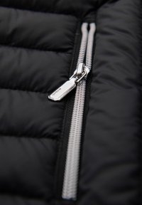 Detail černé péřové bundy ukazuje stříbrný zip. Zip má obdélníkovou táhlo a je obklopen texturovanou látkou.