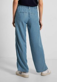 Pantalones de pierna ancha en azul claro con cintura elástica, que cuentan con dos bolsillos traseros, combinados con zapatillas blancas. Textura de denim suave.