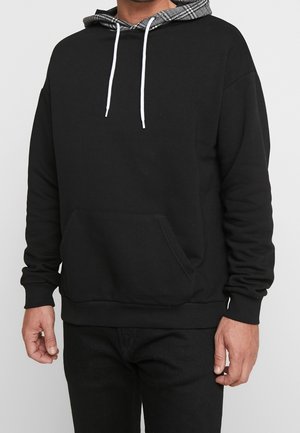 Czarna bluza z kapturem z przednią kieszenią typu kangur oraz białymi sznurkami, noszona z czarnymi spodniami, ukazująca górną część ciała i ręce na jednolitym tle.