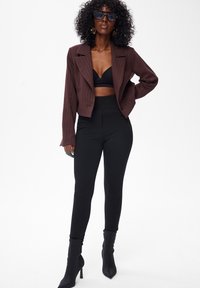 Brązowa marynarka w prążki do talii noszona na czarnym topie biustonoszu, w połączeniu z czarnymi legginsami z wysokim stanem i czarnymi kozakami na wysokim obcasie.