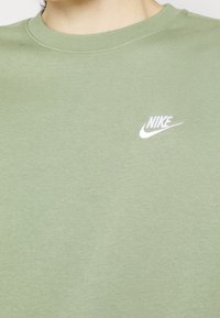 Personne portant un t-shirt Nike vert clair avec un logo Nike blanc sur la poitrine gauche, montrant l'encolure et le haut du torse.