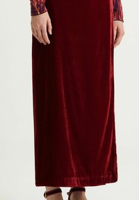 Maxirock aus burgunderrotem Samt, glatte Textur, gerade geschnitten, sichtbarer Saumabschluss, kombiniert mit braunen Riemchensandalen.