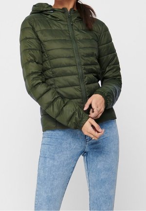 Person iført en olivengrøn quiltet jakke med hætte og lyseblå jeans, justerer jakkeærmerne med begge hænder.