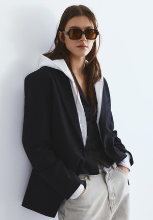 Femeie purtând un sacou negru supradimensionat peste un hanorac gri și un tricou negru, pantaloni bej și ochelari de soare dreptunghiulari maro, pozând cu mâinile în buzunare.