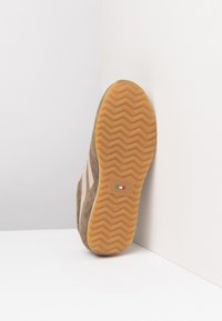 Semelle de basket en daim marron avec un motif en chevrons, présentant des bandes latérales de couleur claire et un logo de marque en vert et rouge.