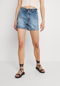 Shorts de denim en azul claro, con un ajuste relajado, cinturilla elástica y dobladillo deshilachado. Combinados con sandalias negras de tiras.