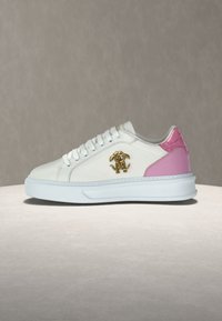 Λευκό αθλητικό παπούτσι με ροζ λεπτομέρεια στη φτέρνα, χρυσό διακοσμητικό λογότυπο στο πλάι, λευκά κορδόνια και παχύ λευκό πέλμα, παρουσιασμένο σε ουδέτερη επιφάνεια.